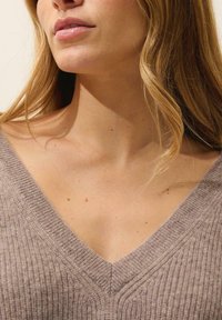 Jasno brązowy sweter w serek o prążkowanej teksturze. Szeroki kołnierz i miękki materiał, odsłaniający delikatnie skórę. Gładkie wykończenie z minimalnymi detalami.