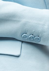 Μανίκι κοστουμιού από υφή υφάσματος σε ανοιχτό γαλάζιο, διπλωμένο προς τα επάνω, δείχνοντας τρία κουμπιά που ταιριάζουν και μια τσέπη με καπάκι από κάτω.