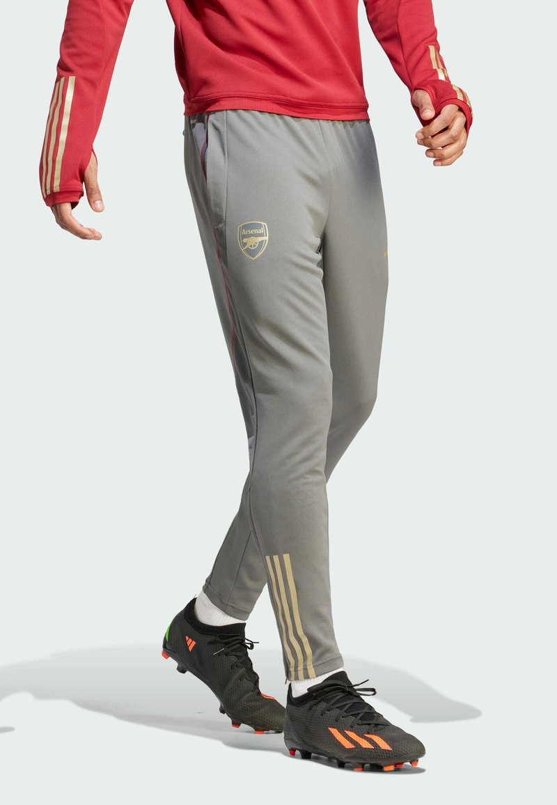 Pantalones deportivos grises con acentos dorados, corte ajustado, que presentan el logo del Arsenal, rayas en los lados y cintura elástica. Calzado negro con detalles en naranja.