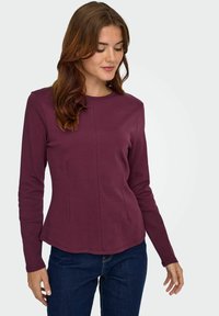 Burgundy langemouwen top van een zachte stof met een ronde halslijn en zijkanten, licht uitlopende zoom, gedragen met donkerblauwe jeans.