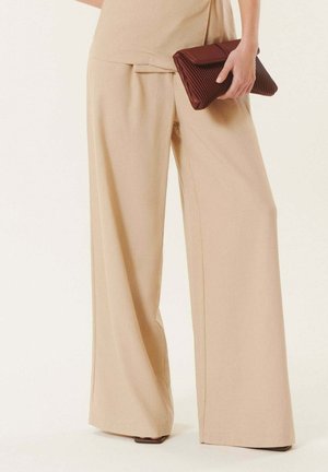 Pantalon beige à jambes larges associé à un haut assorti, tenant une pochette marron pliée à la hanche.
