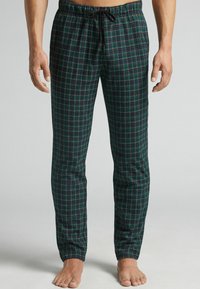 Hombre que lleva pantalones de pijama de cuadros verdes y azul marino con un cordón negro, de pie descalzo frente a un fondo plano.