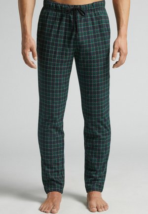 Man in een groen en marineblauw geruit pyjamabroek met een zwarte trekkoord, staand op blote voeten tegen een effen achtergrond.