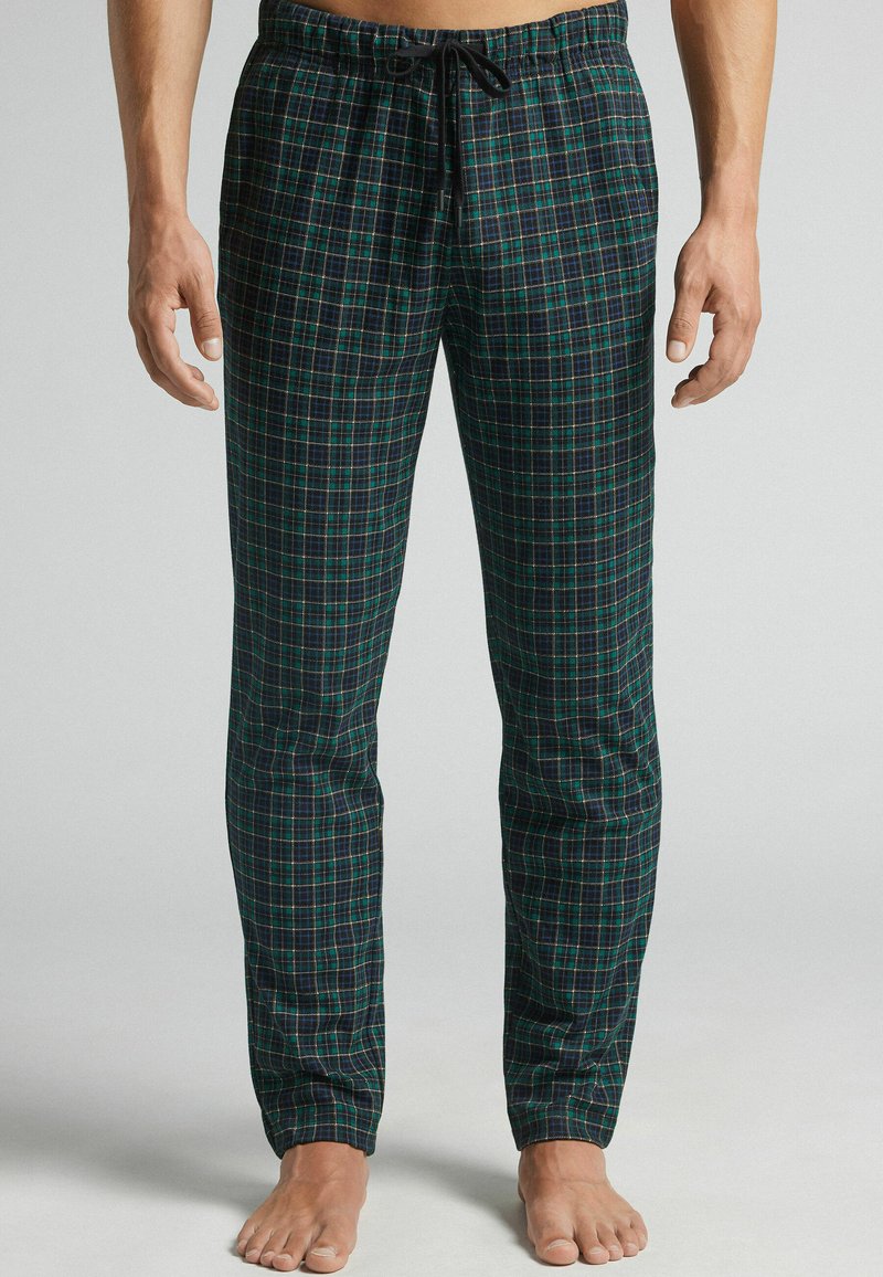 Hombre que lleva pantalones de pijama de cuadros verdes y azul marino con un cordón negro, de pie descalzo frente a un fondo plano.