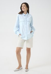 Camicia a righe blu chiaro e bianca con bottoni e due tasche, abbinata a pantaloni corti crema e scarpe basse argentate. Tessuto morbido, vestibilità comoda.