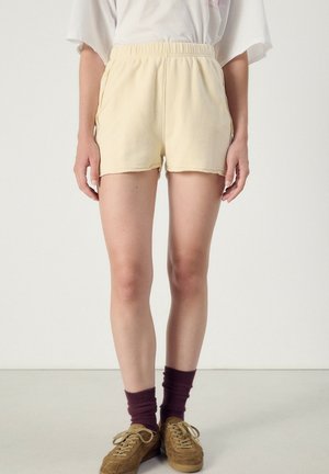 Person wearing light beige elastic waist shorts, brown socks, and brown lace-up shoes, standing against a plain light background.