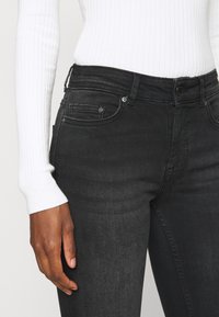 Jeans pretos com um corte slim, apresentando uma cintura de altura média, design de cinco bolsos e detalhes em metal, combinados com um suéter branco com nervuras.