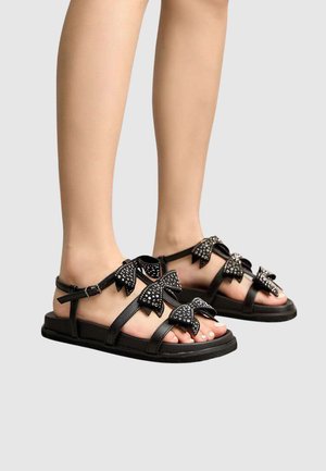 Pieds portant des sandales plates noires avec plusieurs brides en forme de nœud ornées de clous argentés sur un fond clair uni.