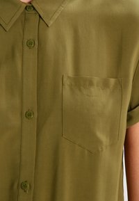 Camisa de manga corta en color verde oliva, hecha de una tela suave, con cuello de punta y un único bolsillo en el pecho. Cierre de botones en la parte frontal.