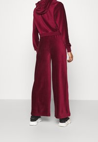 Persona con una sudadera de terciopelo burdeos y pantalones de pierna ancha, de pie con la parte de atrás visible, combinada con zapatillas chunky en blanco y negro.