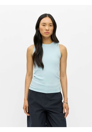 Femme aux longs cheveux foncés portant un haut côtelé sans manches bleu clair et un pantalon large foncé, se tenant devant un fond blanc uni.