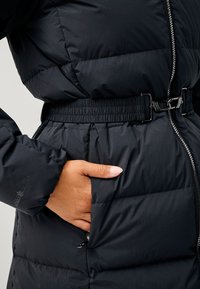 Sort pufferjakke med en indsnøret talje med elastikbånd, lynlåslukning foran og sidelommer. Glat, mat tekstur med minimal detaljer.