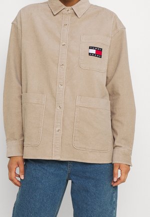 Camisa beige de pana con botones, con bolsillos en el pecho y en la parte inferior frontal, con un parche del logo de Tommy Jeans en el bolsillo del pecho.