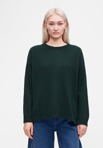 Jeune personne aux longs cheveux blonds lisses, portant un pull vert foncé à col rond et un jean bleu, debout devant un fond clair uni.