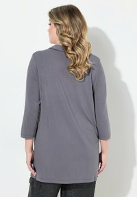 Tunic grigia a maniche lunghe con colletto arrotondato, fessure laterali e tessuto morbido. La parte posteriore presenta un design semplice senza dettagli prominenti.