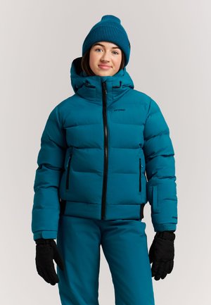 Jonge persoon die een teal winterjas draagt, bijpassende broek, teal gebreide muts en zwarte handschoenen, staand tegen een effen lichte achtergrond.