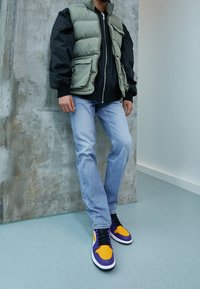 Person som står inomhus iklädd ljusblå jeans, lila och orange sneakers, svart jacka och grön vadderad väst mot betong- och vitmålade väggar.