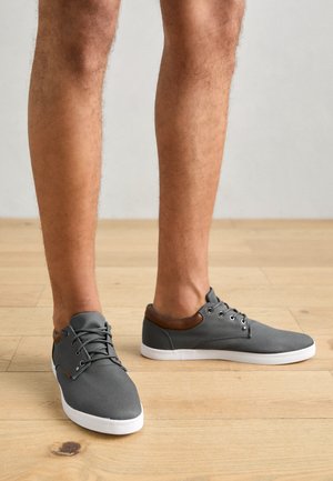 Szürke vászon sportcipők bőrbetéttel a bokánál, fehér gumi talppal, kerek orral és fűzős dizájnnal. Fapadló háttér.