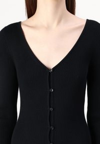 Cardigan noir à rayures, à manches longues, avec un col en V et six boutons noirs sur le devant. Présente un design ajusté et un tissu texturé.