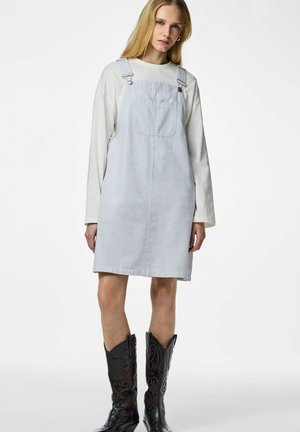 Femme aux longs cheveux blonds portant une chemise blanche à manches longues, une robe tablier rayée bleu clair et des bottes de cowboy noires brodées.