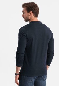 Man med brunt hår som bär en marinblå långärmad tröja och blå jeans, visad bakifrån mot en enkel vit bakgrund.