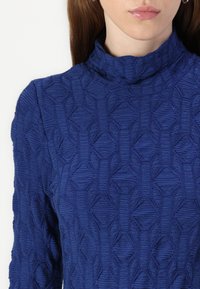 Top albastru cu mânecă lungă și guler înalt. Materialul texturat prezintă un model geometric de linii și forme ridicate care creează adâncime.