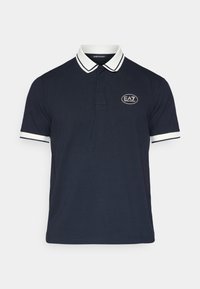 Μπλε πουκάμισο πόλο ναυτικού χρώματος με λευκή ρίγα στο γιακά, κοντά μανίκια και λογότυπο κεντημένο σε οβάλ σχήμα στην αριστερή πλευρά του στήθους.