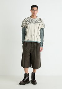 T-shirt à manches courtes, crème, avec des motifs abstraits noirs superposés sur une chemise à manches longues bleue, associé à un short à carreaux marron et des bottes noires.