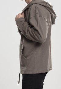 Persona che indossa una giacca con cappuccio grigia, con tasche frontali, che tiene il colletto, mostrata di profilo su uno sfondo neutro.