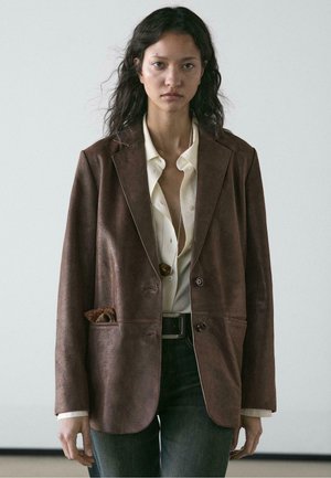 Brauner Lederblazer mit strukturiertem Design, zwei Außentaschen und Knopfverschluss. Kombiniert mit einer weißen Bluse und Jeans.