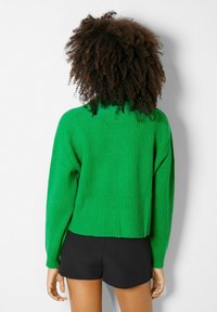 Zelený žebrovaný úpletový svetr s vysokým límcem a spadlými rameny, doplněný černými šortkami. Textura je měkká a robustní.