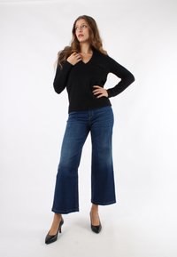 Maglione nero a scollo a V, aderente in vita, abbinato a jeans svasati blu scuro e tacchi alti neri, su uno sfondo bianco semplice.