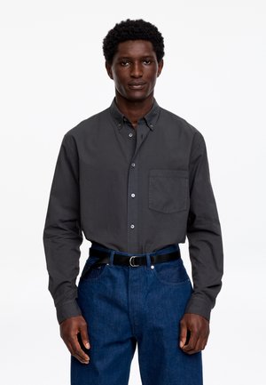 Mężczyzna ubrany w ciemnoszarą koszulę na guziki, wpuszczoną w niebieskie dżinsy z czarnym paskiem, stojący na tle białego tła.