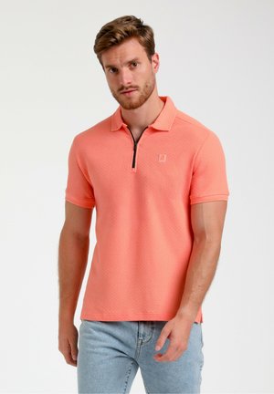 Polo à manches courtes en tissu corail texturé, avec col zippé, couleur unie, et un petit logo sur la poitrine, associé à un jean bleu clair.