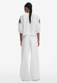 Blusa blanca corta con mangas abullonadas que presenta bordados florales azules; combinada con pantalones anchos blancos, con una textura suave en toda su extensión.