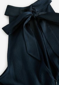Mörkblå topp med en texturerad väv och knutdetalj vid halsen samt en flytande design. Materialet verkar lätt och mjukt.