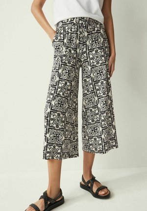 Mujer con pantalones anchos para la pierna con estampado en blanco y negro, top blanco sin mangas y sandalias negras con tachuelas, de pie con las manos en los bolsillos.