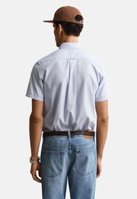 Homme portant une chemise bleu clair à manches courtes avec boutons, ceinture tressée marron, jean bleu clair, casquette marron, dos tourné vers un fond uni.