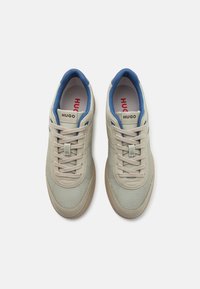 Baskets beiges avec une tige texturée et des accents en cuir lisse, doublure bleue et semelle extérieure en caoutchouc marron clair. Elles présentent des lacets beiges et des détails de logo.