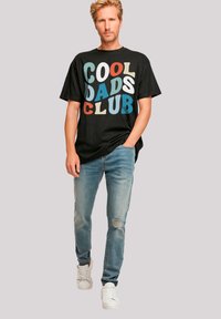 Black cotton t-shirt with multicolored "COOL DADS CLUB" graphic, paired with light blue distressed jeans and white sneakers.