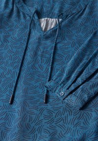 Blå mönstrad skjorta med V-ringning, som har subtila svarta vågmönster, dragsnoddar och knäppta ärmslut i ett mjukt tyg.