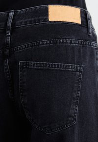 Svarta denimjeans med en läderlapp vid midjan med präglad text, en bakficka och synliga sömdetaljer.