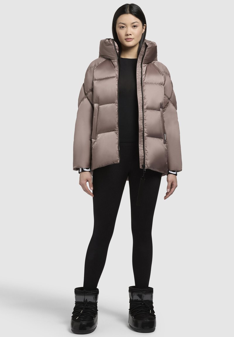 Giacca puffa di colore marrone chiaro con colletto alto, zip frontale e grandi sezioni trapuntate. Indossata con leggings neri e stivali neri lucidi.
