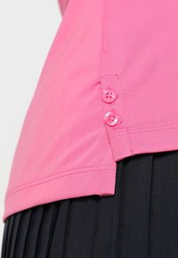 Růžová košile s krátkým rukávem, která má hladkou texturu, boční rozparek a dva růžové knoflíky jako akcent, v kombinaci s černou plisovanou sukní.