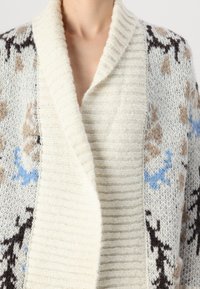 Πλεκτό κάρντιγκαν με κρεμ ριμπ σακάκι, και μοτίβο στο σώμα σε ανοιχτό μπλε, καφέ και μαύρο σε μπλε φόντο.