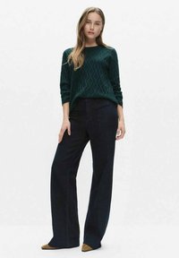 Donkergroene, gestructureerde trui met een ronde halslijn, gecombineerd met zwart, hooggetailleerde, wijd uitlopende broek en puntige bruine schoenen met blokhak.