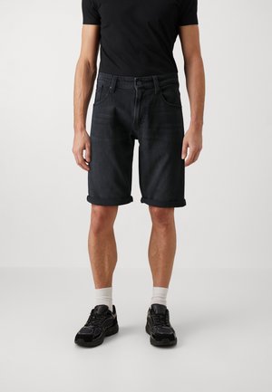 Homme portant un short en jean noir retroussé, un t-shirt noir, des chaussettes blanches et des baskets noires, debout devant un fond clair uni.
