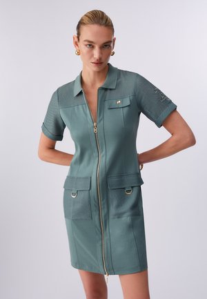 Donna con i capelli pettinati all'indietro che indossa un vestito verde acqua con zip, maniche corte, tasche con patta e dettagli in oro con bottoni e anelli.