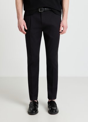 Uomo che indossa pantaloni neri slim fit con dettaglio piega, abbinati a scarpe eleganti in pelle nera e cintura nera.