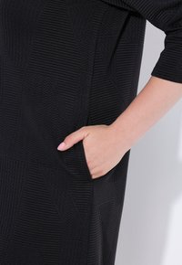 Abito nero con tessuto testurizzato, caratterizzato da motivi geometrici e una tasca laterale. Maniche lunghe e vestibilità comoda.